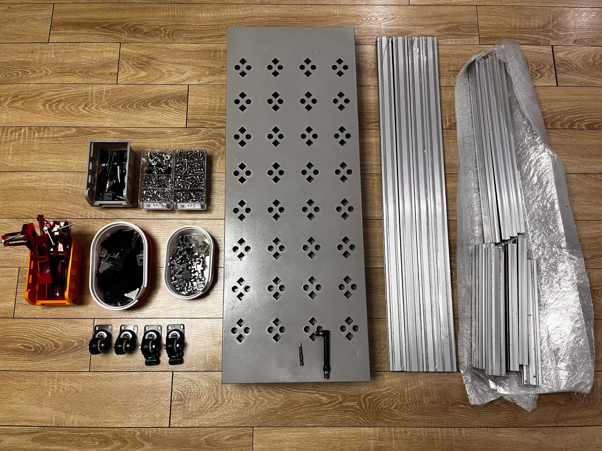 2040 aluminum extrusion pieces, perforated metal sheets, hardware components, and trolley wheels laid out on wooden floor