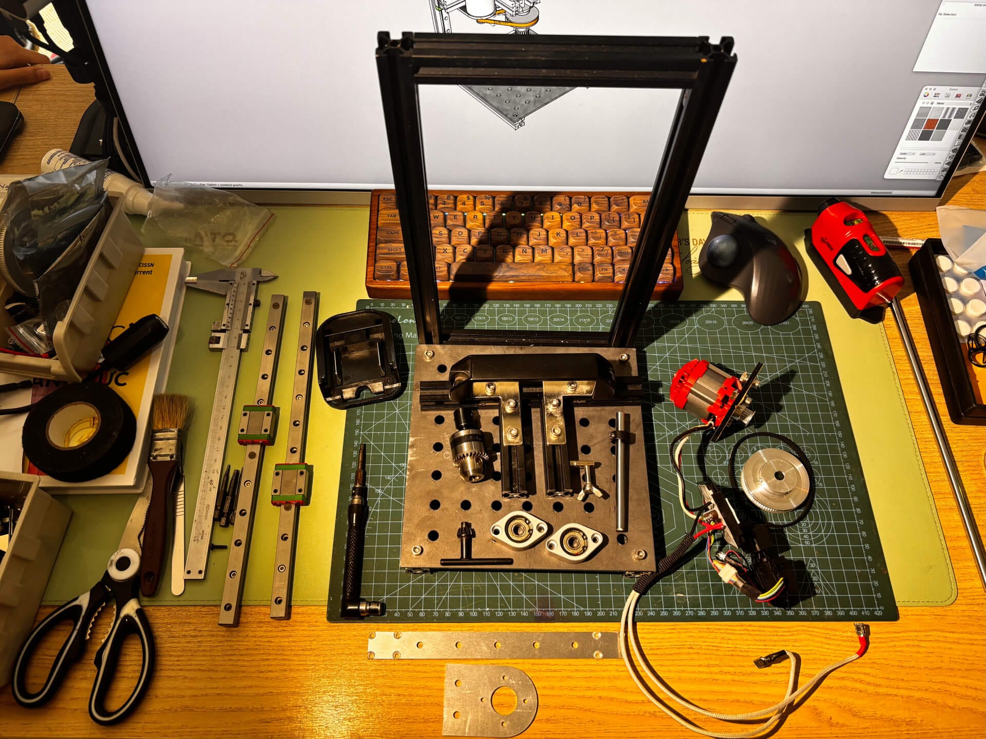 Top-down view of press drill components laid out on a cutting mat including aluminum extrusion frame, linear rails, brushless motor, and assorted hardware