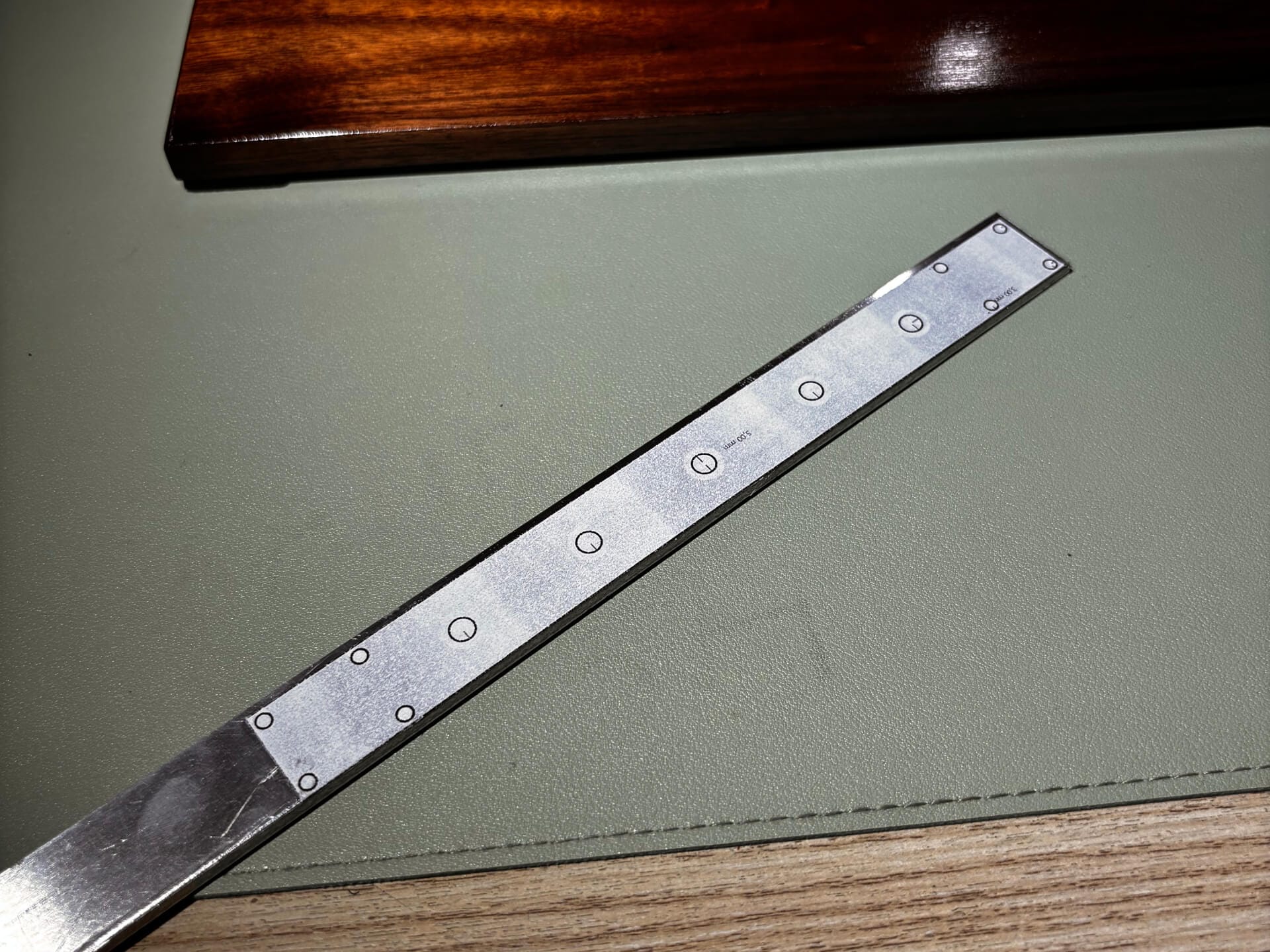 Aluminum flat bar with marked and drilled holes laid on desk mat with wooden keyboard in background
